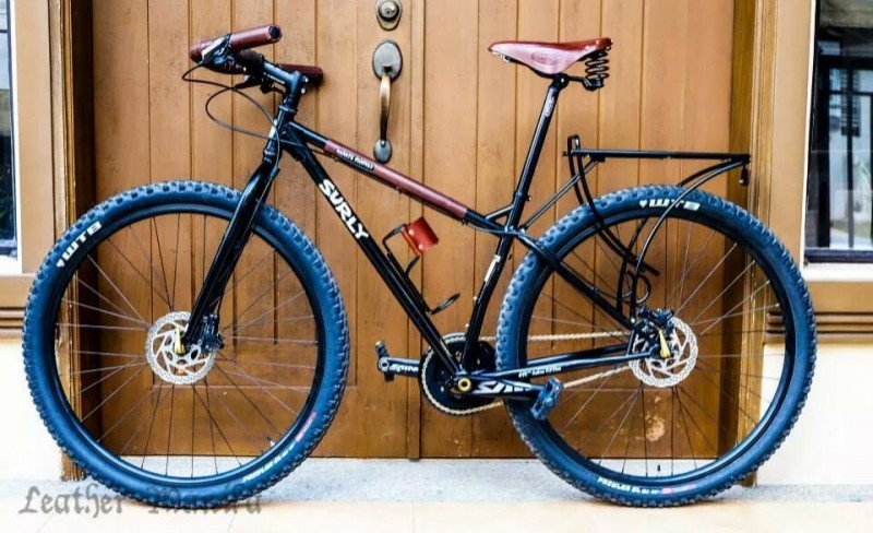 Left side view of a black Surly Karate Monkey bike with leather grips and seat, parked in front of 2 brown, wood doors