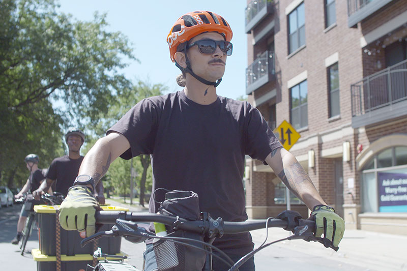 Person wearing bike helmet, sunglasses, stopped to deliver food on Surly Big Easy cargo bike with trailer