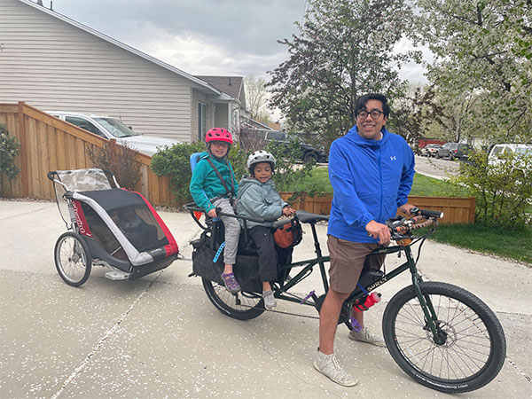 Abner standing over Big Easy with two kids on back of bike and two kids in bike trailer behind, in driveway