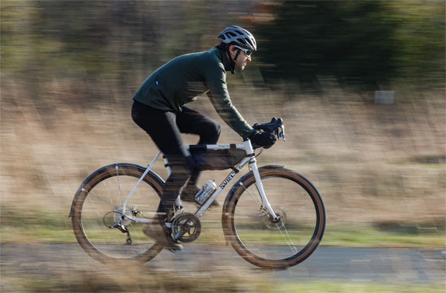 Right side view of a cyclist on a Surly Midnight Special bike speeding down a paved trail with tall brush shown behind