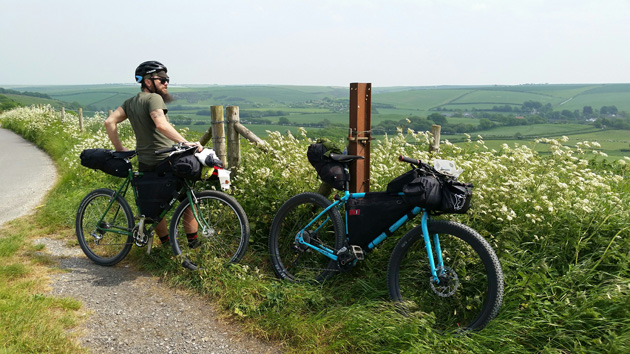 A cyclist in the grass on the side of the road standing with their bike, next to a bike with no rider leaning on a fence