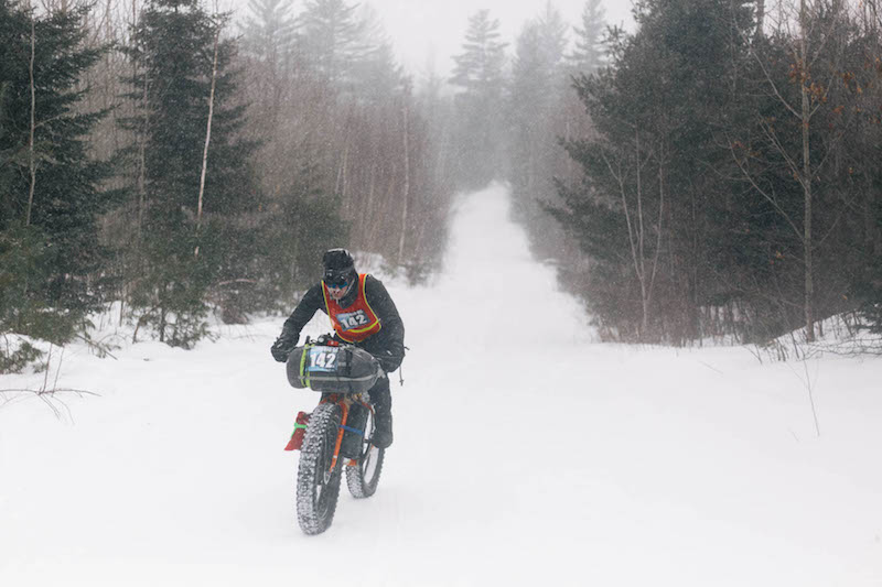 Front view of a cyclist riding a fat bike up a snow covered road hill in the forest on a snowy day