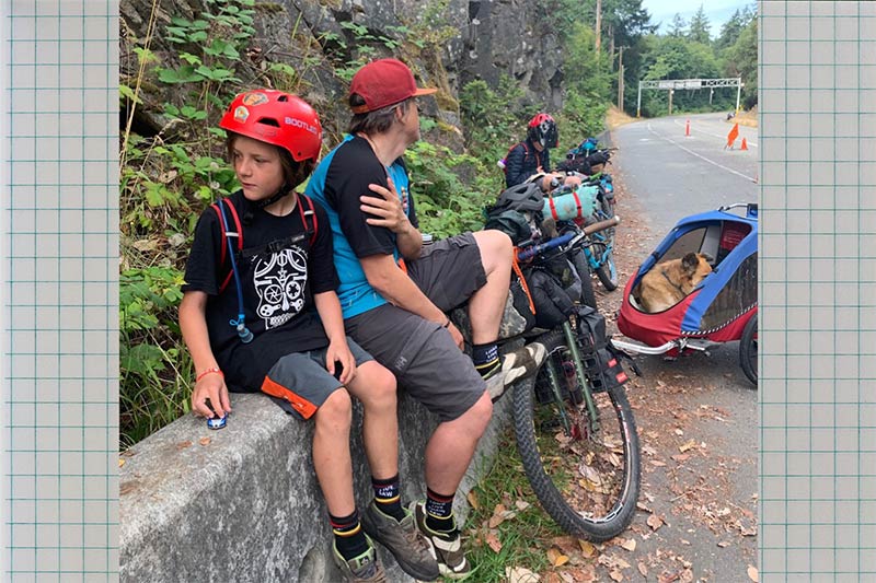 Sitting on concrete wall with loaded bikes and trailer with dog parked family takes a break on side of road