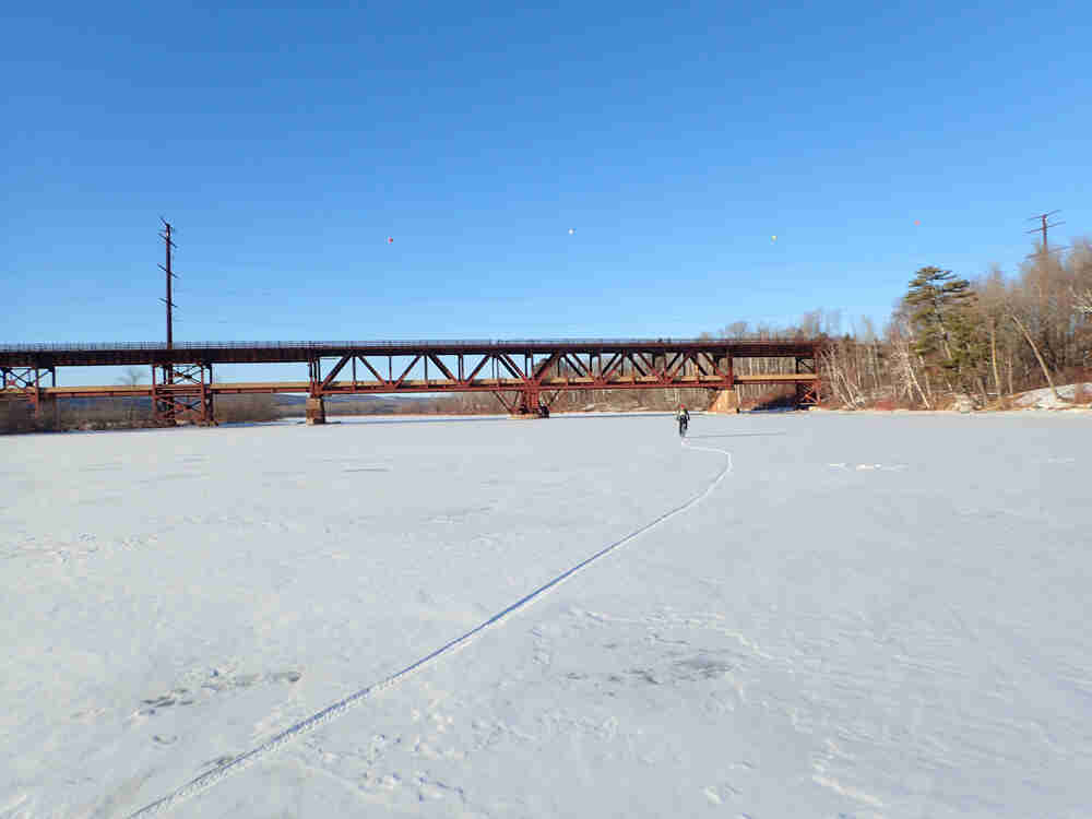 A cyclist riding a bike on a frozen river towards a bridge, leaving a trail in the snow