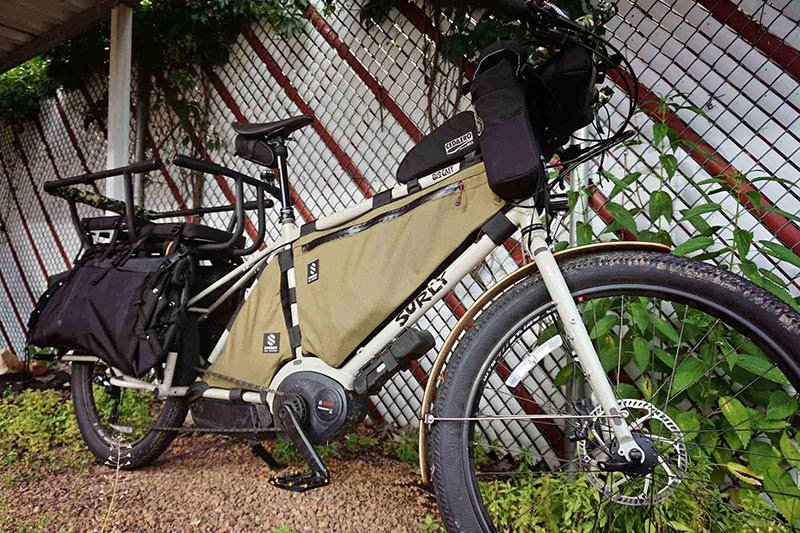 Left side view of a white Surly Big Easy bike with packs and racks leans on a chain link fence underneath a deck