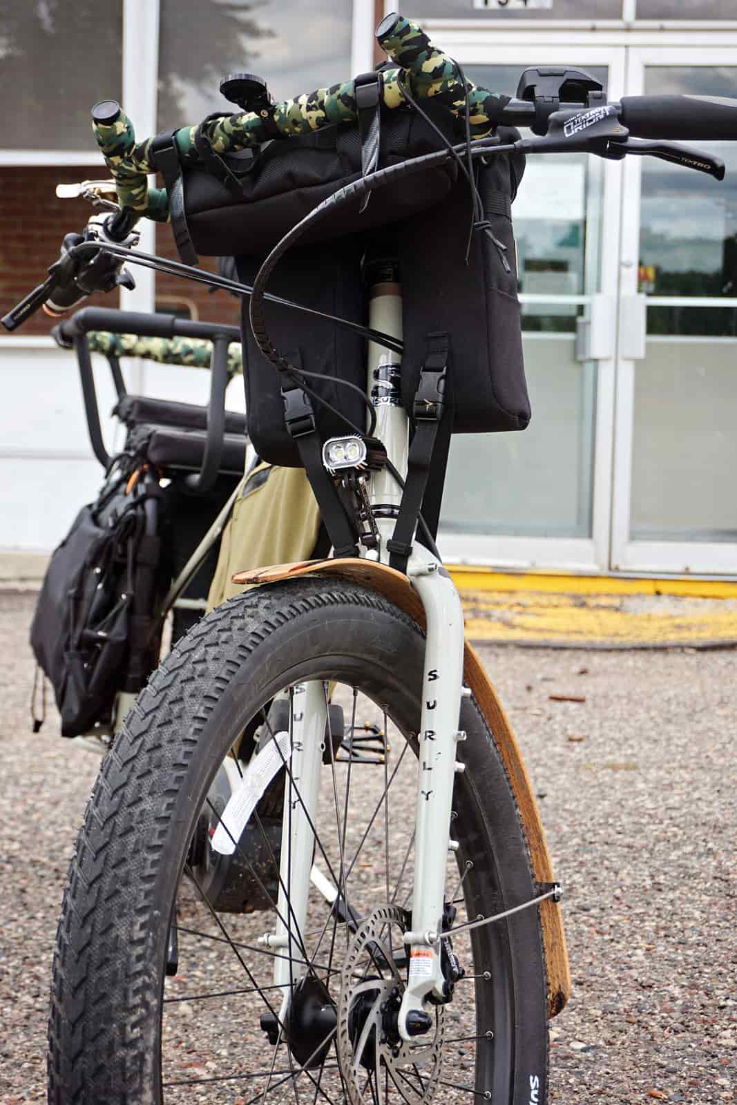 Front view of a white Surly Big Easy bike with gear and headlight on pavement in front of a building with glass doors