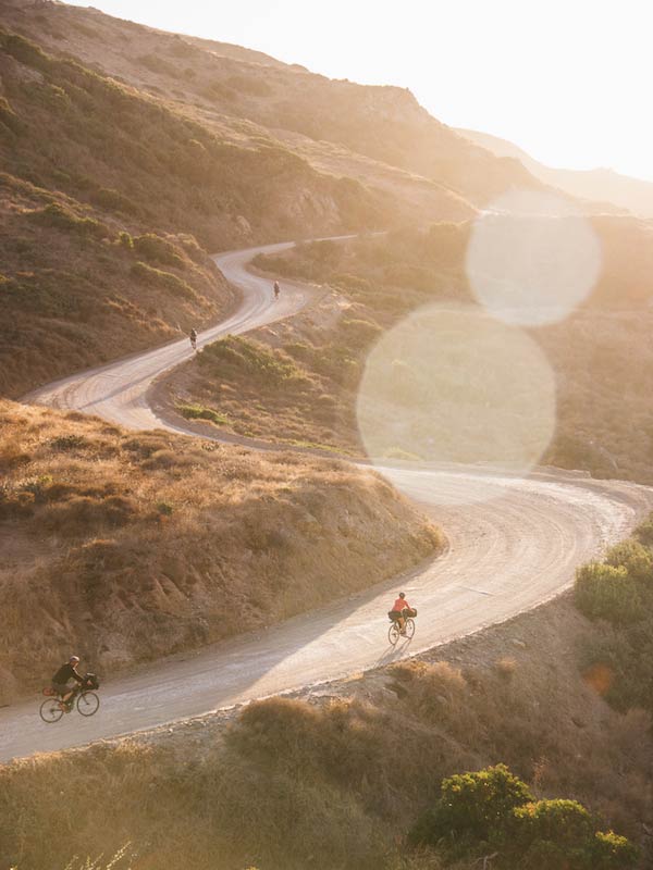 4 cyclist riding up a windy gravel road in grassy, bushy hills with bright sunshine above