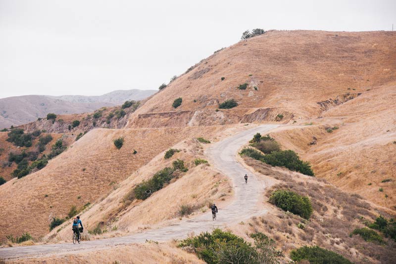 Rear view of 3 cyclist riding up a windy gravel hill towards the side of a brown, grassy mountain hill