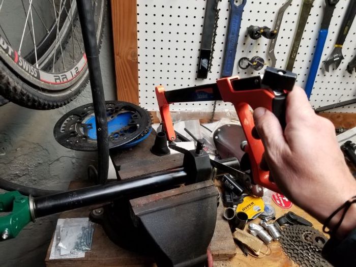 A right hand holding a hacksaw on a fork tube mounted in a vise with tools and bike parts on a workbench