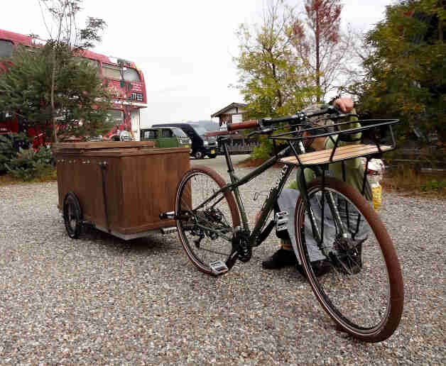 Front right side view of a Surly bike with a wood trailer hitched behind, with a person squatting near the front