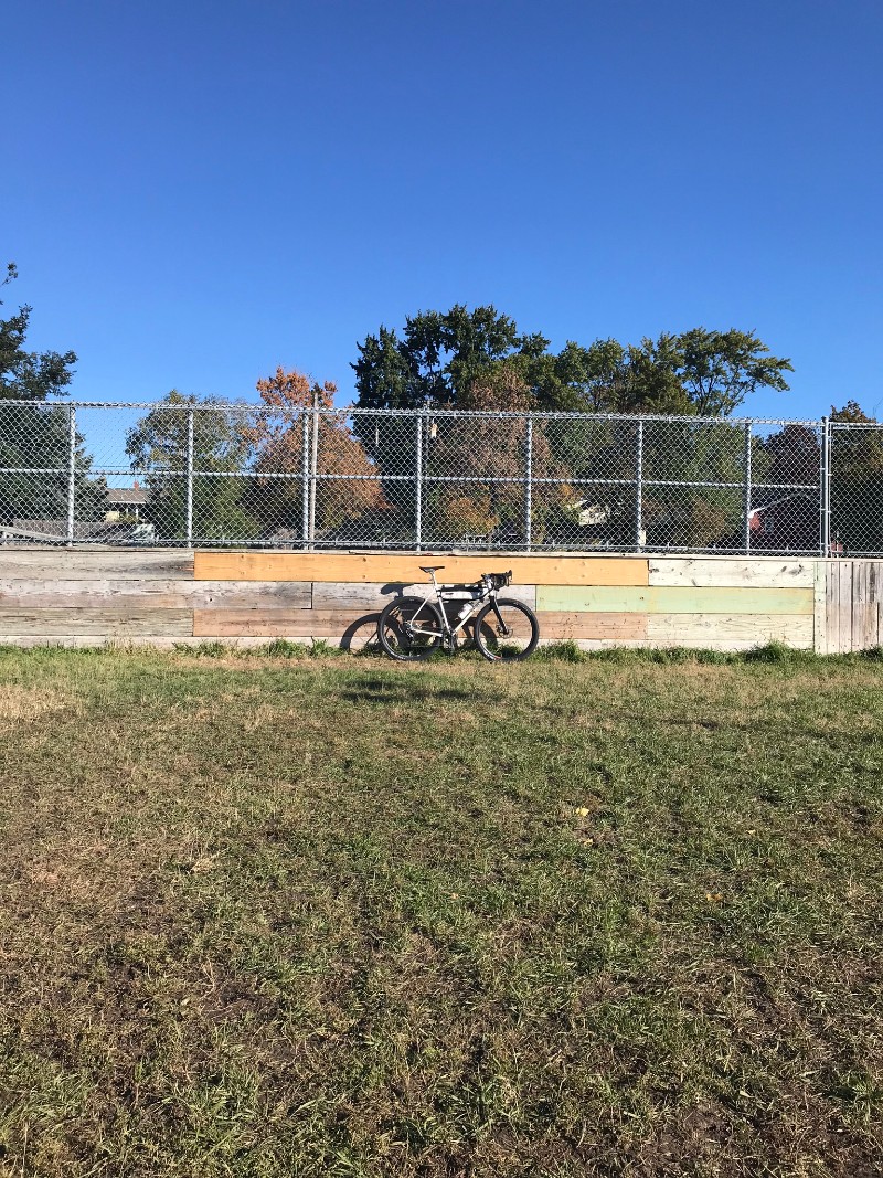 Surly Midnight Special Bike on grass leaning on a board wall with chain link above in a park, trees in the background