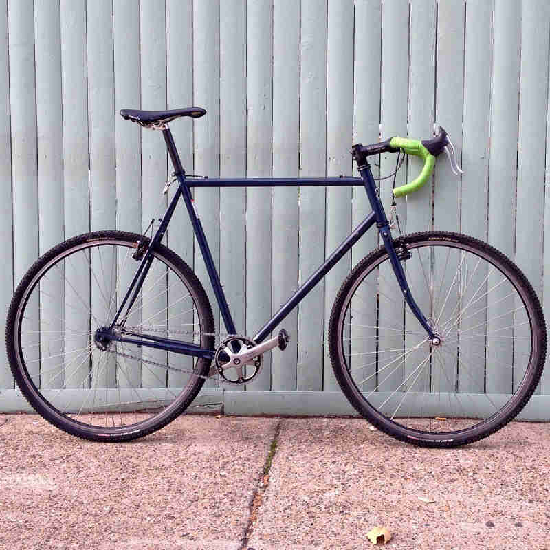 Right side view of a blue Surly Cross Check bike, parked on a sidewalk along a steel fence