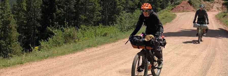 Cyclist smiles riding up a hill on a gravel road in the pine trees to the side with another rider tailing behind