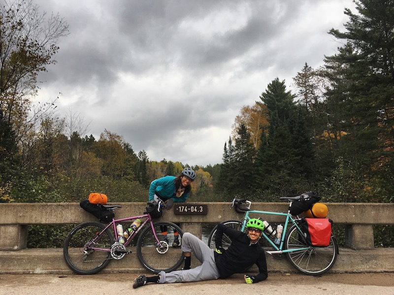 Front view of two cyclists posing on a concrete bridge with trees behind, on a cloudy day