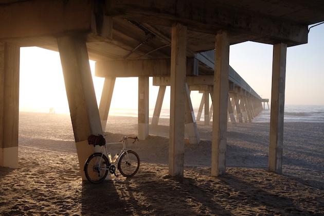 Surly Midnight Special bike leaning on a support in the sand under a pier with the sun shining through