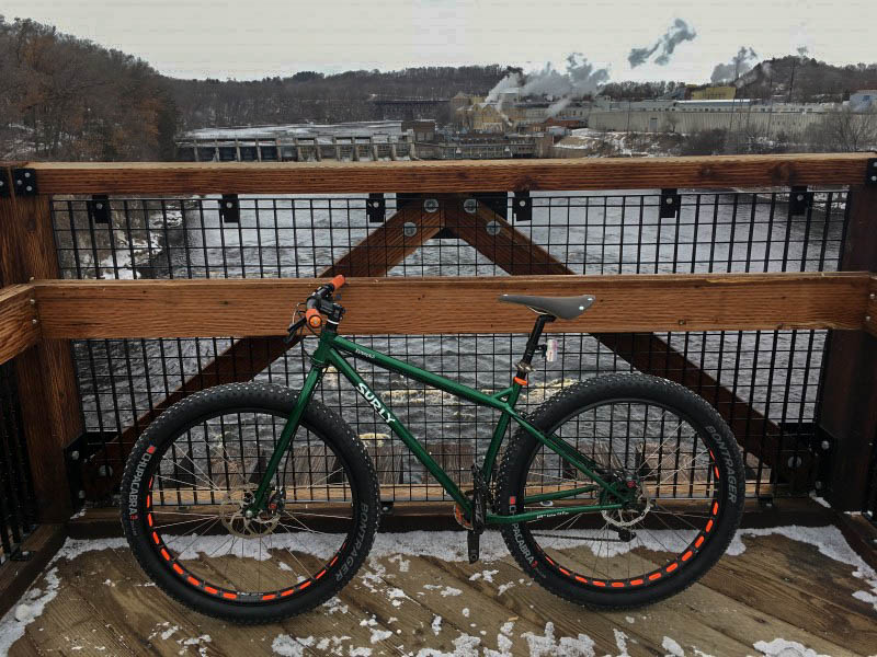 Left profile of a green Surly fat bike on a bridge over a river, with a dam in the background