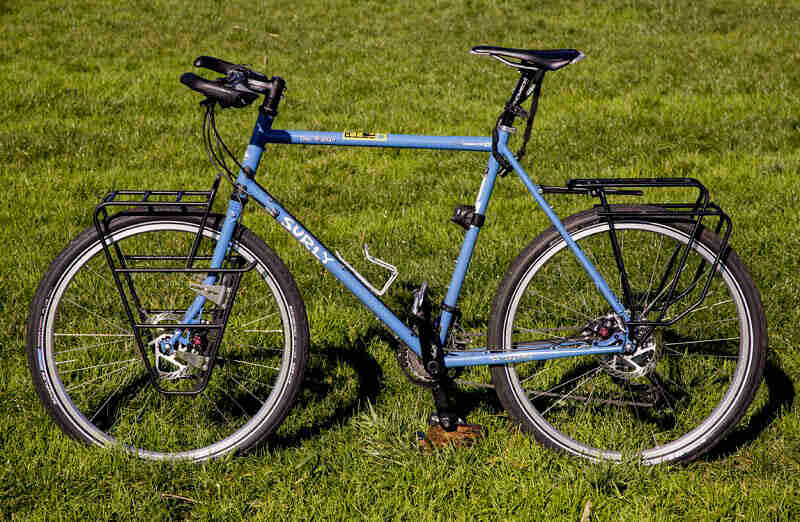 Left profile of a blue Surly bike with front and rear racks, standing in a green, grass field