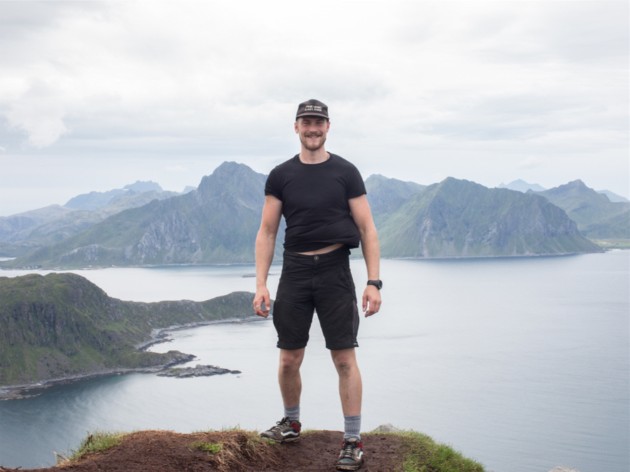 Jac Mac standing on a rock cliff above lake with mountains in the background