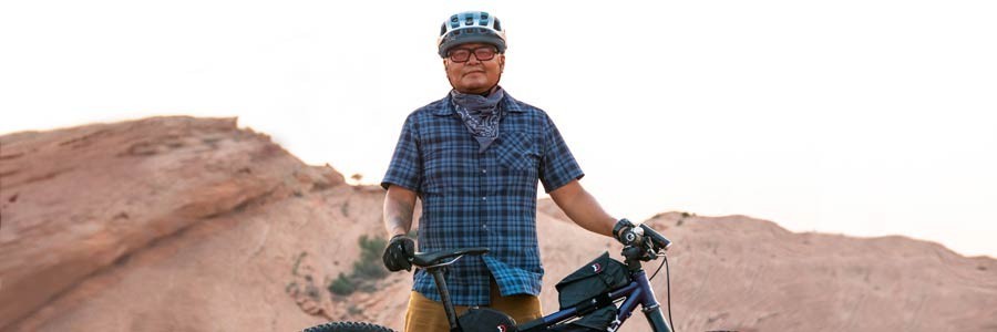 Jon Yazzie standing with loaded mountain bike wearing cycling helmet and riding glasses in front of slick rock