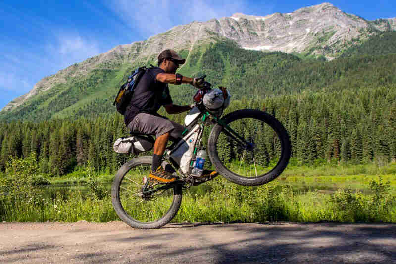 A cyclist riding a wheelie on a Surly Krampus bike, green, on a paved road, with trees and mountains in the background