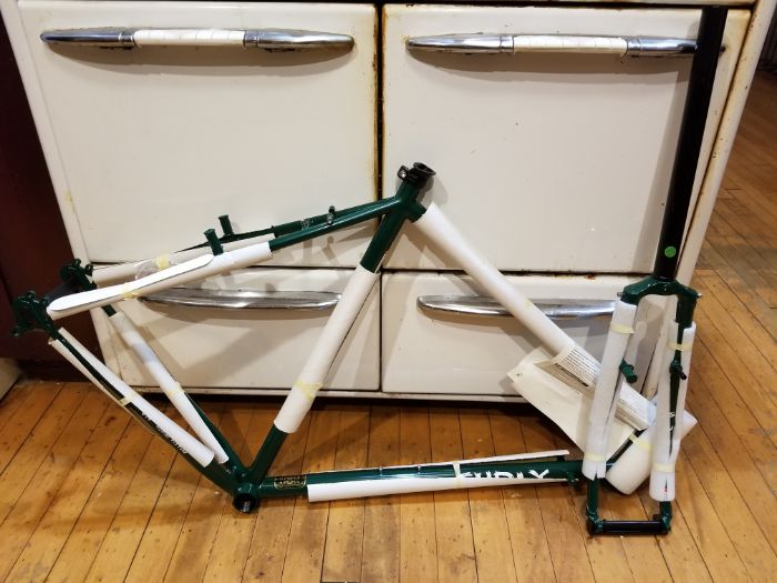 Right side view of a wrapped Green Surly Pack Rat frame and fork leaning against a white 4 draw cabinet on a wood floor