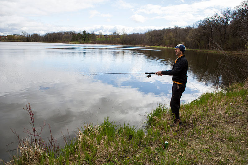 Person in warm clothing and cycling cap fishing from grassy lake shore on cloudy day