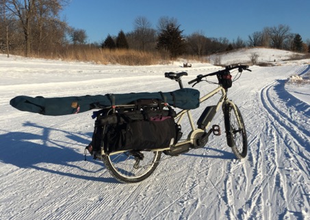 Surly Big Easy bike, tan, with rear saddle and ski bag shown on top of a snow covered cross country ski trail in nature