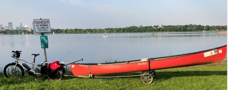 Left side view of a tan Surly Big Easy bike with a trailer carrying a red canoe attached, in the grass beside a lake