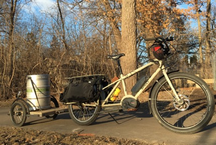 Right profile view of a tan Surly Big Easy bike with gear pulling a trailer with keg on a paved trail with trees behind