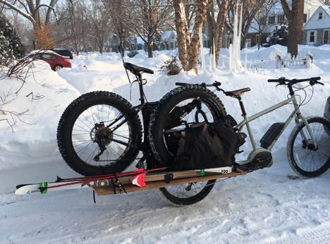 Right side view of a tan Surly Big Easy bike and trailer hauling a fat bike and skis on a snow in a neighborhood