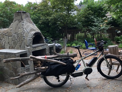 Right side view of a tan Surly Big Easy bike, with saddle bags loaded with firewood, next to an outdoor wood stove