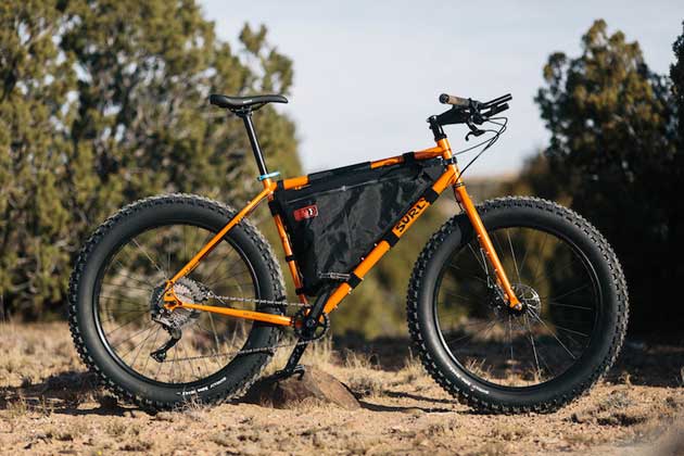 Right side view of an Surly Pugsley fat bike, orange, with inner frame pack, on sandy and rocks with trees in the background