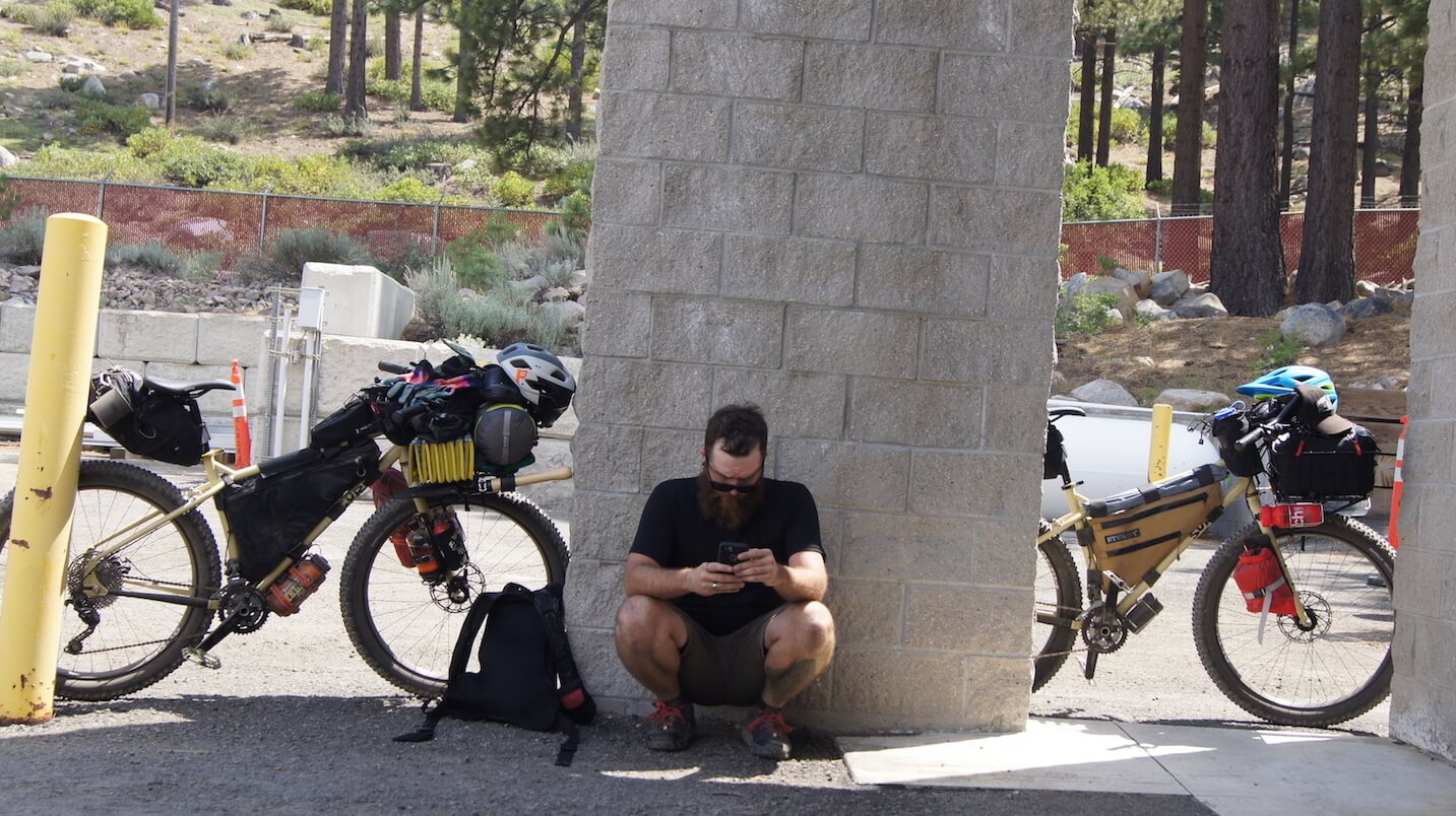 Front view of a squatting against a brick pillar, with of bikes, loaded with gear, at the sides