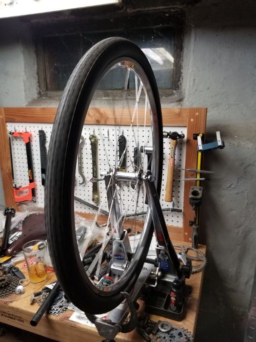 Assembled bike wheel with tire mounted to a rim truing device on a workbench with pegboard with tools and bike parts