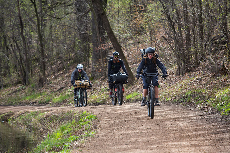 Cyclists wearing warm clothing riding bikes loaded with gear on gravel forest road next to water