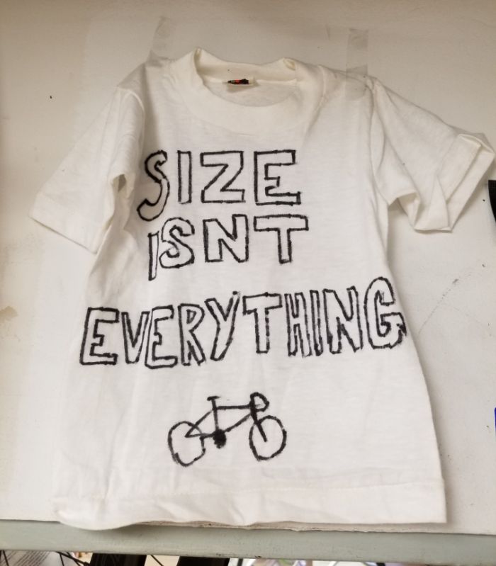 White T-Shirt with the words 'size isn't everything' and a picture of a bike is taped to a white wall