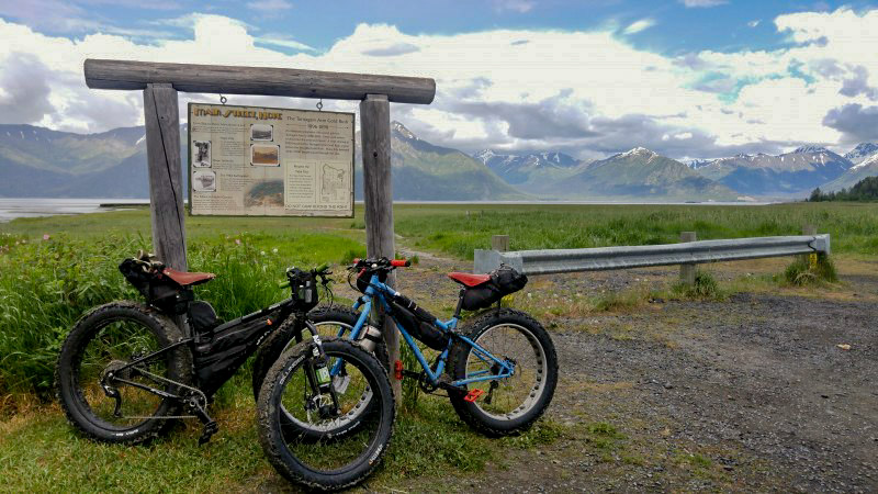 Side view of 2 Surly Ice Cream Truck fat bike, parked on gravel, in front of a sign, with a grassy field behind