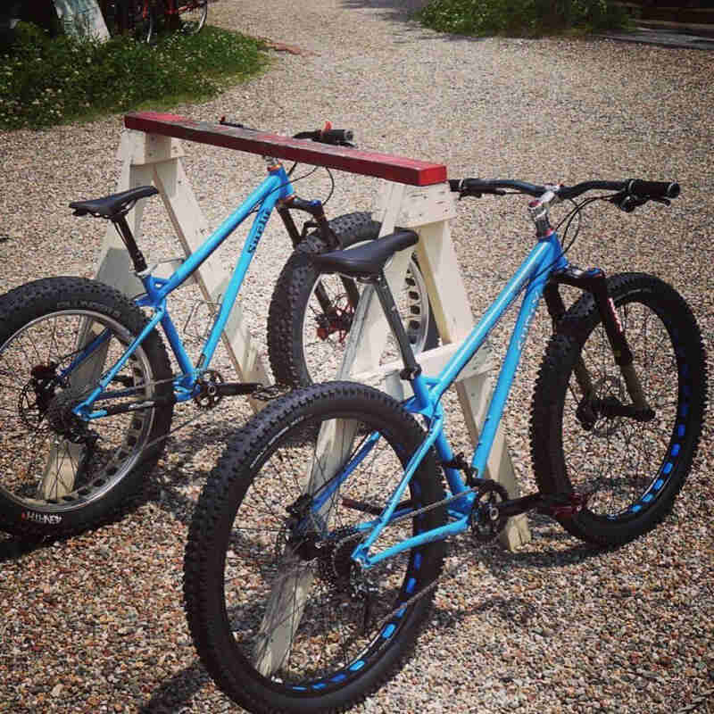 Right rear angle view of 2 Surly Ice Cream Truck fat bikes, parking on gravel, leaning against a sawhorse