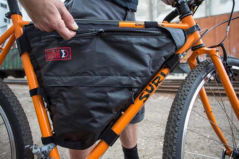 Side view of black frame pack installed on orange Surly bike and person unzipping pocket on frame pack