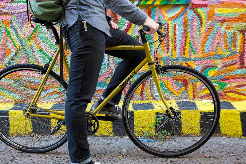 Cyclist in casual clothing standing over yellow Surly bike with one foot on pedal and one foot on ground in front of mural