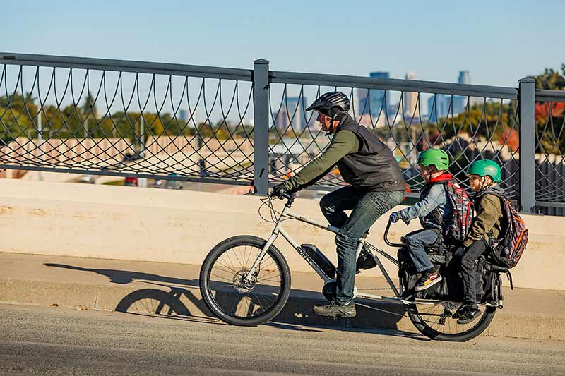 Cyclist riding Surly Big Easy cargo ebike with two children on back over highway bridge with skyscrapers in background