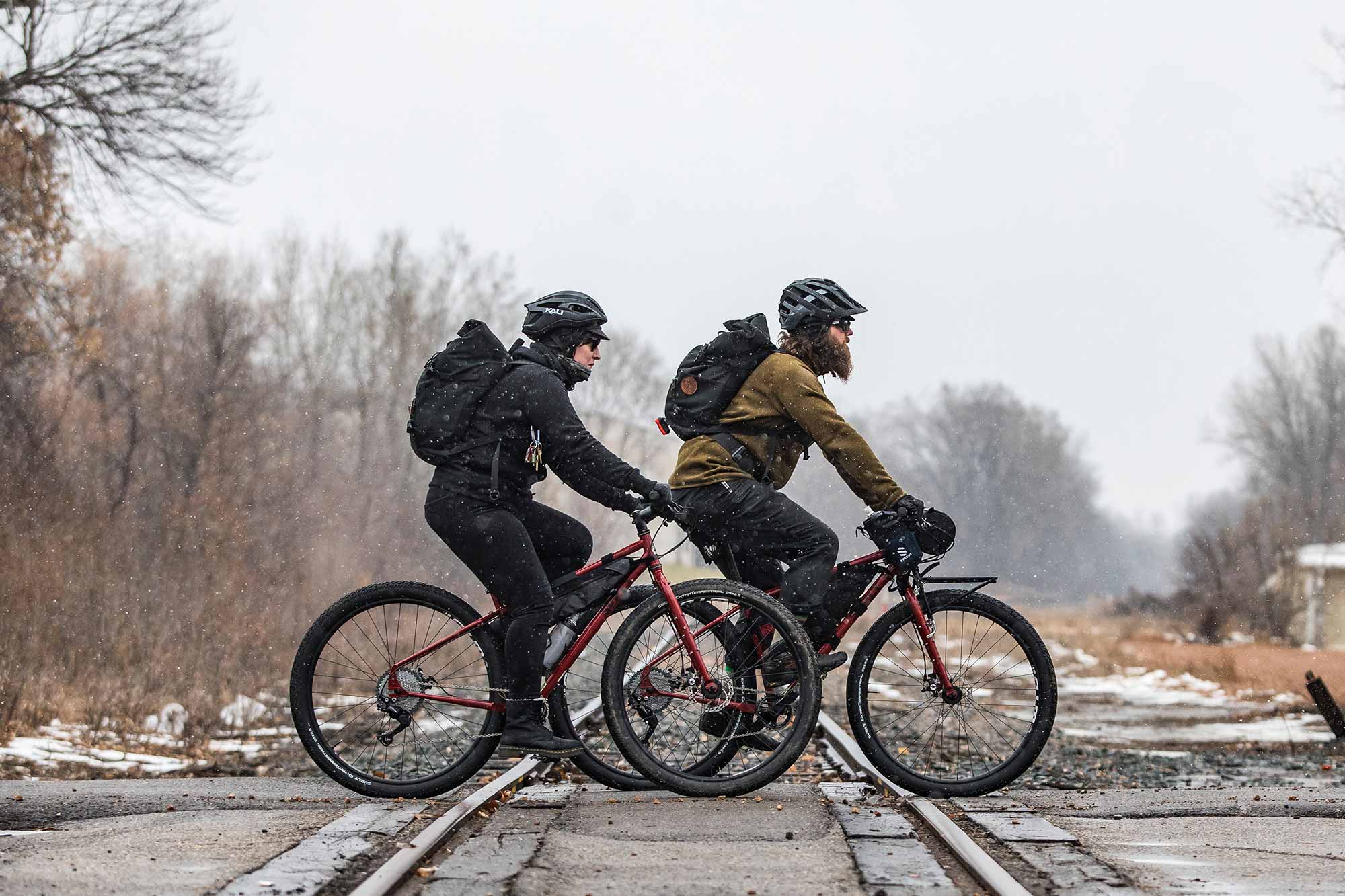 Two bike commuters riding on road in warm clothing, helmets and backpacks in light snow