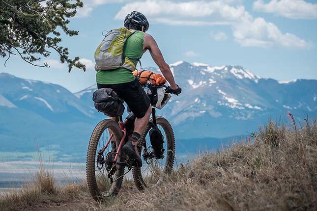 Mountain biker with helmet, backpack and bikepacking gear attached to bike on trail with mountains in background