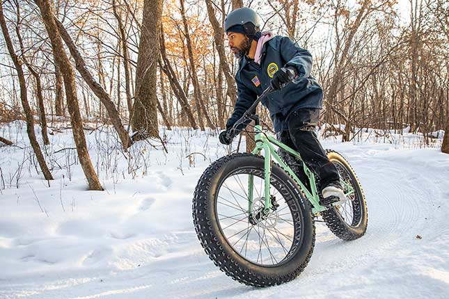 Mountain biker wearing helmet and warm clothing riding mint colored Surly Fat Bike on snow covered off-road trail