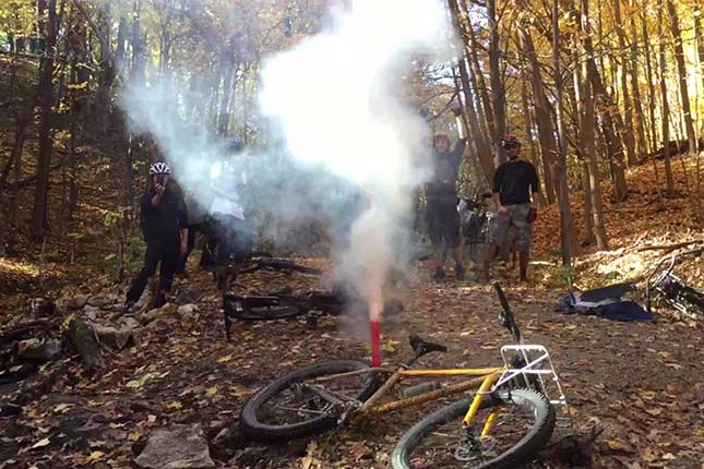 Group of bikers in woods standing around with bikes laying on ground with smoke erupting from red firework tube