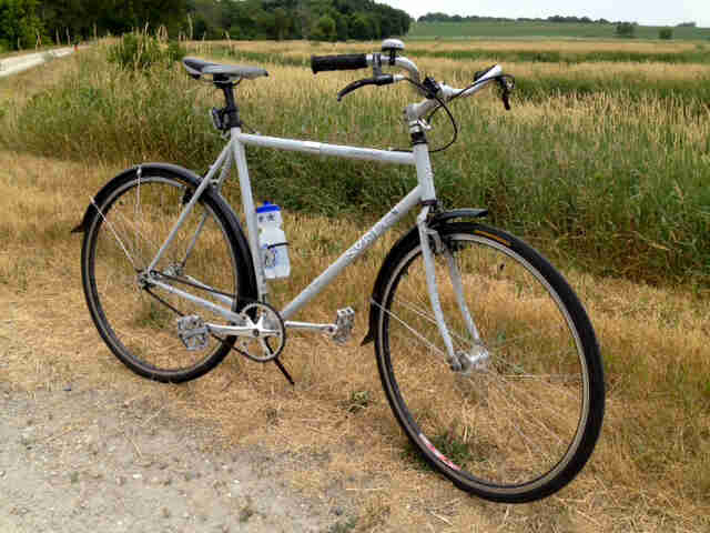 Right side view of a white Surly Cross Check bike, parked next to a gravel road, with a grassy field in the background