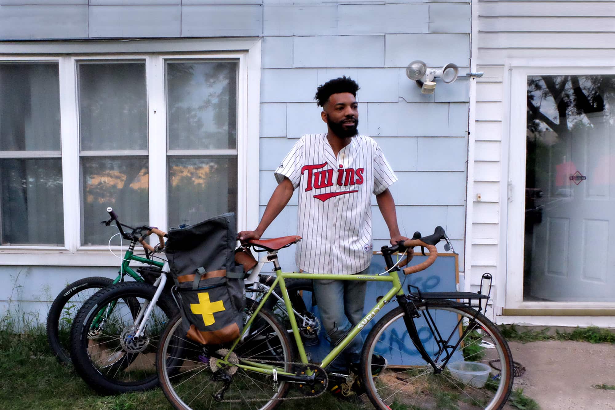 Wesley stands in front of house wearing Twins jersey with lime color Surly bike in front of him and 2 bikes behind him