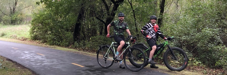 Two cyclists stand on a paved trail with their Surly bikes at a right profile view, green leafed trees in woods behind