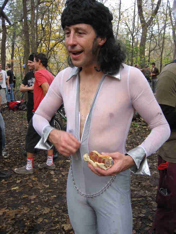 Front view of a person wearing a black wig and wearing a white bike race suit, with people and trees in the background