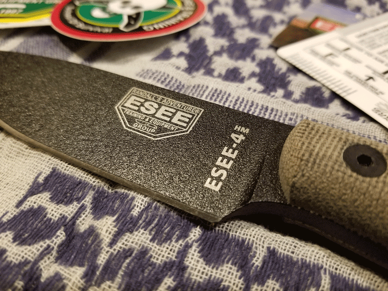 ESEE 4HM knife with black powder coated and showing small portion of a tan handle lying on woven blue and white blanket 